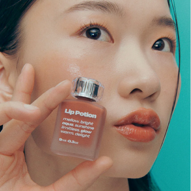 Model holding Alternative Stereo Aqua Glow – warm brown glossy lip tint with radiant finish.