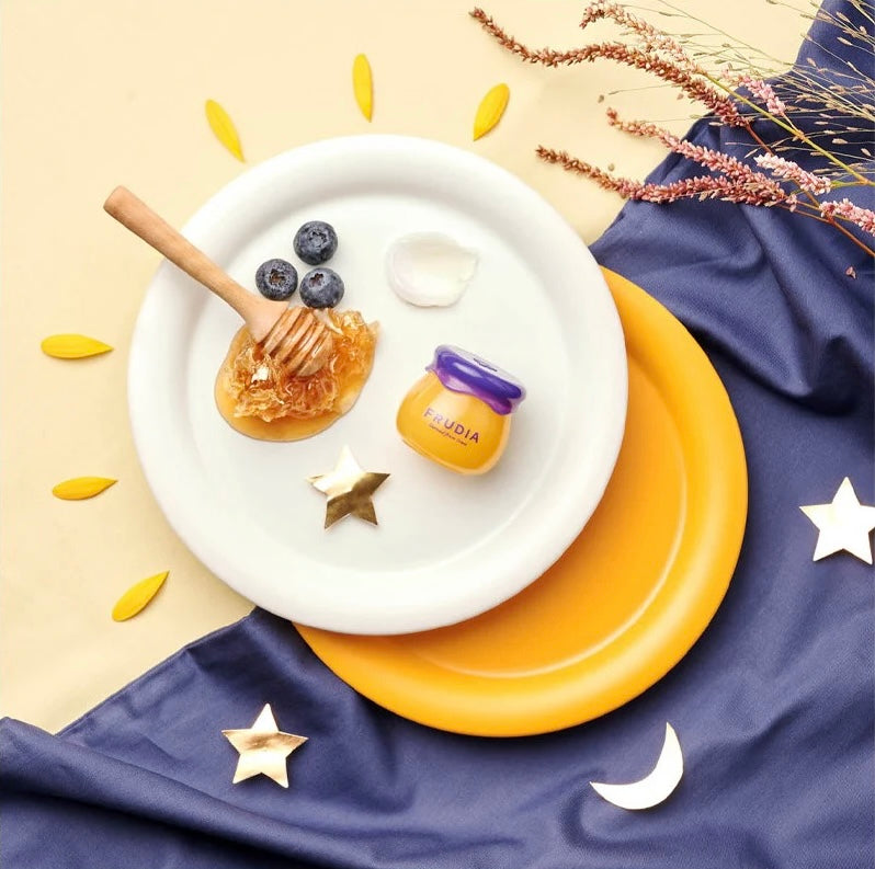 Frudia lip balm styled with honey, blueberries, and decorative plate setup.