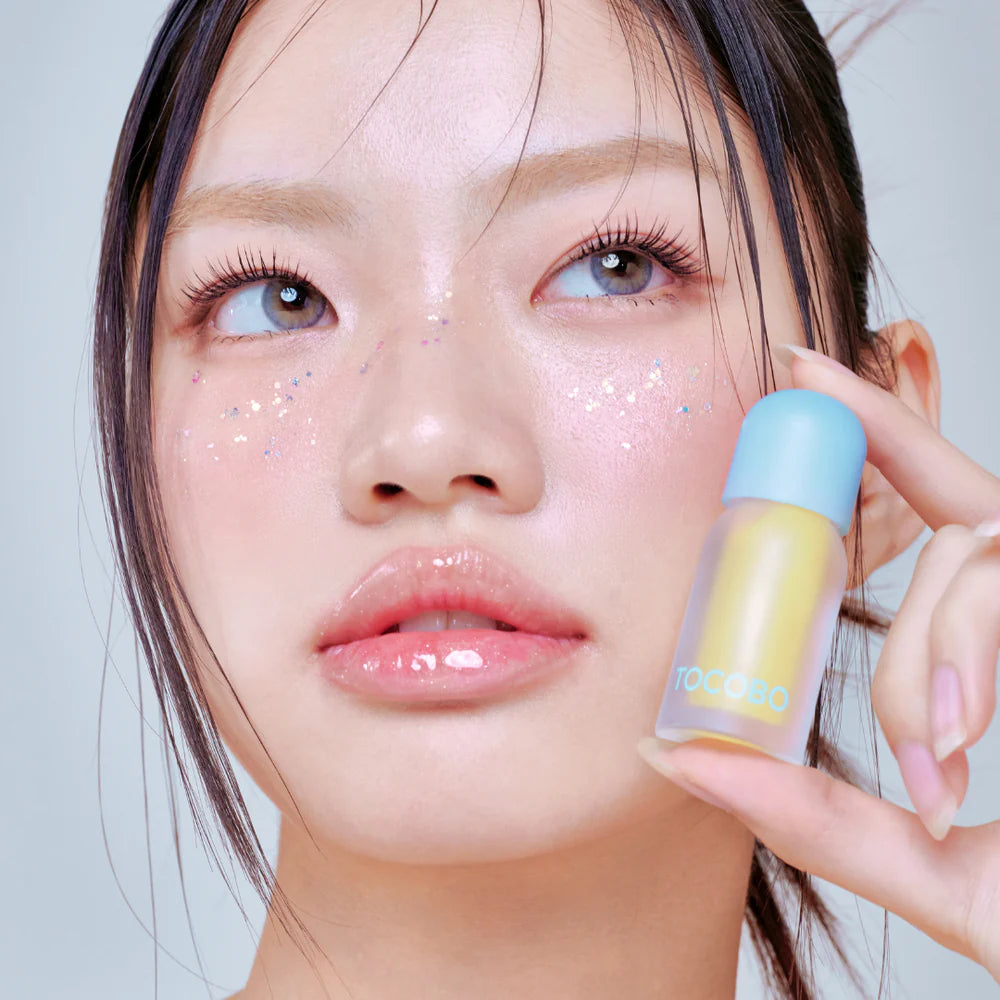 Model with sparkly makeup holding yellow TOCOBO lip oil.