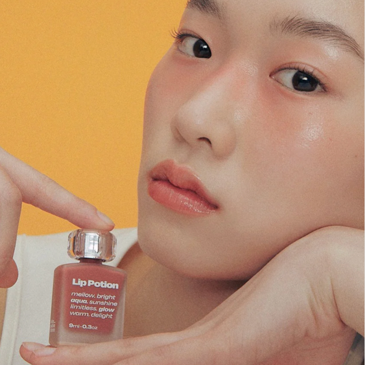 Alternative Stereo Aqua Glow lip tint held against orange background – warm glow, natural finish.